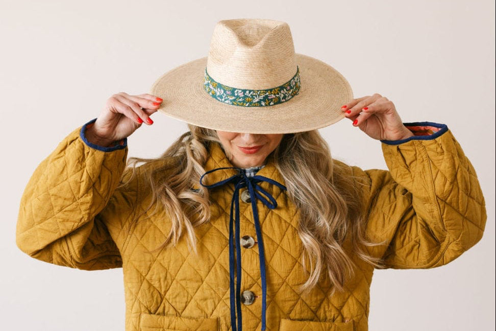 Person wearing a mustard yellow quilted jacket and a wide-brimmed hat with a floral band on a plain background